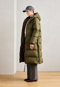 Maslinasto zeleni puffer kaput s kapuljačom, sa ušivenim dezenom, dugom dužinom i bočnim detaljima od zatvarača, uparen s sivim širokim trapericama i smeškim čizmama.