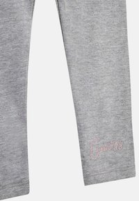 Szürke pamut leggings, amelyek puha textúrával rendelkeznek. A bal lábon egy rózsaszín hímzett "Guess" logó látható a szegély közelében.