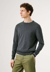 Suéter de punto gris con cuello redondo y puños acanalados, combinado con pantalones verde oliva. Presenta detalles en negro en contraste en la manga.