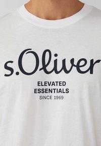 Witte katoenen T-shirt met korte mouwen, voorzien van zwarte tekst: "s.Oliver," "ELEVATED ESSENTIALS," en "SINCE 1969" gecentreerd op de voorkant.