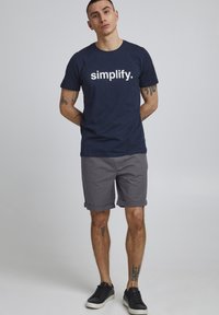 Marineblå bomull T-skjorte med hvit "simplify." tekst, satt sammen med rullede grå shorts og svarte joggesko, som viser et avslappet antrekk.