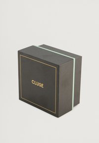 Μαύρο κουτί με υφή, χρυσές και μέντας πράσινες λεπτομέρειες, και ετικέτα "CLUSE" μπροστά. Ορθογώνιο σχήμα με καπάκι.