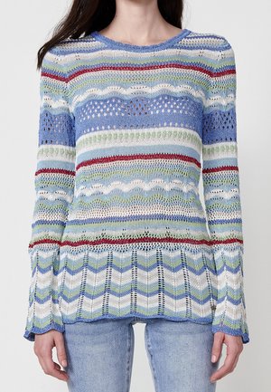 Kobieta w swetrze z długim rękawem o otwartym splocie z poziomymi paskami w kolorze niebieskim, białym, zielonym i czerwonym oraz w jasnoniebieskich dżinsach.