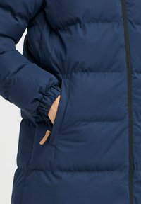 Temno moder podložen plašč z teksturirano tkanino, elastičnimi manšeti in stranskim žepom z bež usnjeno oblogo.