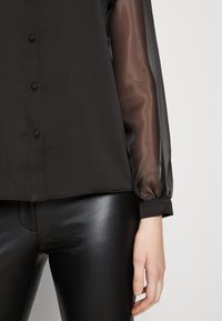 Svart topp med genomskinliga långa ärmar, knappp Detalj och solid kropp, kombinerad med glänsande svarta leggings som betonar textur och kontrast.