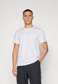 Camiseta blanca de manga corta en tejido suave con un cuello redondo. Presenta un pequeño logo azul de "Peak Performance" en el pecho.