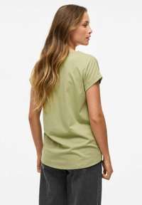 Femme aux longs cheveux ondulés portant une chemise à manches courtes vert clair et un pantalon gris foncé, de dos contre un fond blanc uni.