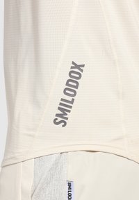 Maglietta sportiva color beige chiaro con tessuto testurizzato. Presenta il logo "SMILODOX" in grigio, con dettagli di cucitura sottili vicino all'orlo.
