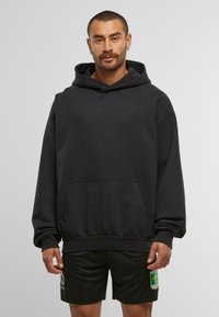 Mann in schwarzem Kapuzenpullover mit Fronttasche und schwarzen Shorts mit gelbem und grünem Emblem am rechten Bein vor einfarbigem Hintergrund.