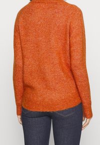 Pomarańczowy wełniany sweter z wysokim kołnierzem, teksturowanym wykończeniem i luźnym krojem. Dolny brzeg jest lekko zakrzywiony. Noszony z ciemnymi dżinsami.