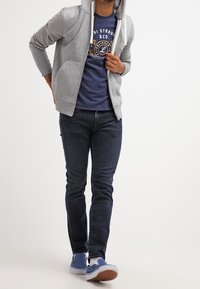 Felpa con cappuccio grigia con cerniera e tasche, indossata sopra una t-shirt grafica blu navy, abbinata a jeans scuri e sneaker blu. Outfit casual con texture stratificate.