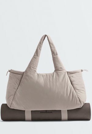 Sac de yoga rembourré beige avec des poignées larges, reposant sur un tapis de yoga gris foncé roulé, sécurisé par des sangles beiges.