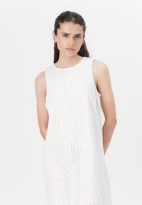 Robe sans manches blanche faite en tissu à œillets, présentant un motif géométrique avec des découpes. Design simple avec un col rond et une coupe décontractée.