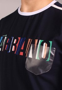 Černé tričko s krátkým rukávem, zdobené vícebarevným texturovaným písmem a průhlednou kapsou. Kontrastní žebrovaný výstřih.