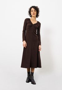 Braunes Langarmkleid mit gerafftem Vorderteil, V-Ausschnitt und fließendem A-Linien-Rock. Das Modell trägt schwarze Knöchelstiefel. Glatte Stofftextur.