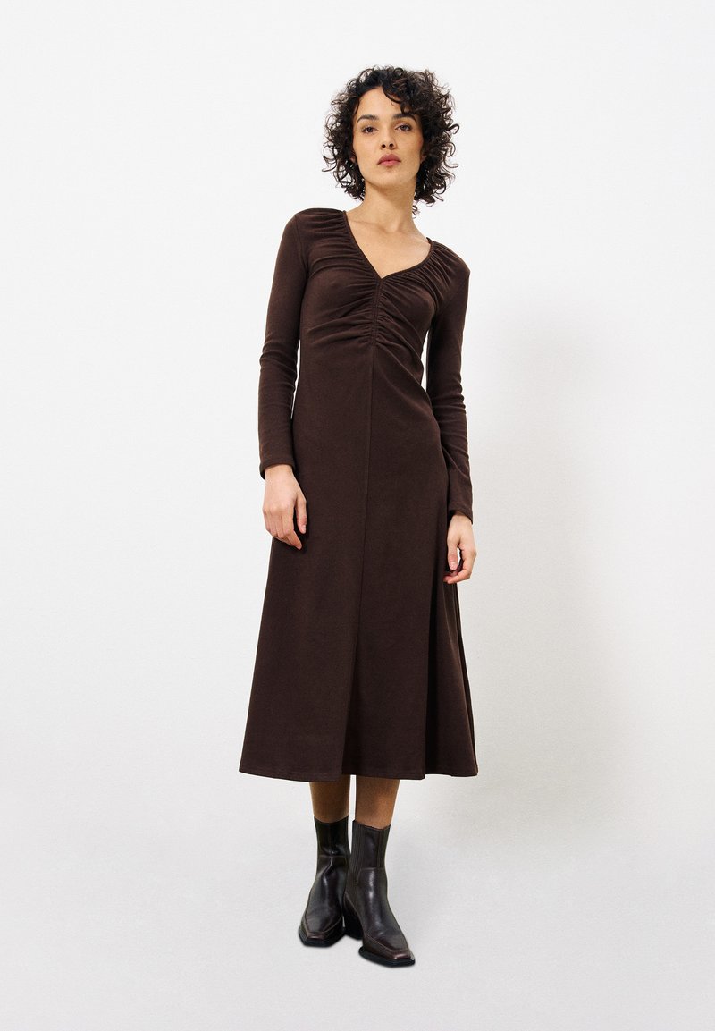 Braunes Langarmkleid mit gerafftem Vorderteil, V-Ausschnitt und fließendem A-Linien-Rock. Das Modell trägt schwarze Knöchelstiefel. Glatte Stofftextur.