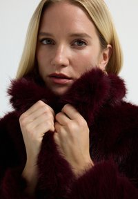 Vaaleahiuksinen nainen pitää syvän burgundinvärisen keinoturkishaalarin kaulusta lähellä kasvojaan, neutraali tausta, pehmeä valaistus korostaa ihon rakennetta.