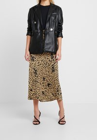 Blazer doppiopetto in pelle nera con vestibilità rilassata, abbinato a una gonna midi con stampa leopardata nei toni beige e neri, completato da tacchi neri con laccetti.