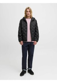 Schwarze gesteppte Daunenjacke mit Kapuze, getragen über einem hellrosa Hemd. Dunkelblaue Jeans und schwarze Schuhe runden das Outfit ab.