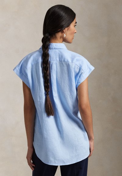 Sieviete ar gariem pinumiem, tērpusies gaiši zilā īss piedurkņu kreklā, redzama no mugurpuses pret neitrālu fonu.