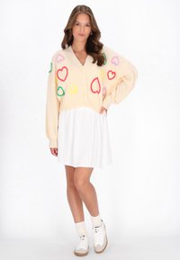 Sweter kardigan w kolorze kremowym z kolorowymi wzorami serc, z zapięciem na guziki, z przesadnie dużymi rękawami, zestawiony z plisowaną białą spódnicą i sneakersami.