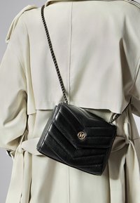 Borsa a tracolla nera in pelle con design a rombi trapuntati, hardware in oro con logo e tracolla in catena nera, indossata sopra un leggero trench.