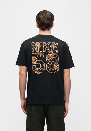 Mies, jolla on päällään musta T-paita, jossa suuri "NIKE 58" -teksti selässä ja oransseilla kasvi- ja geometrisillä kuvioilla koristeltu.