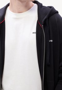 Černá mikina na zip s červenými doplňky přes bílé tričko. Mikina má texturovanou látku, viditelné švy a vyšívané logo.