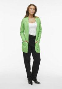 Grön stickad cardigan med öppen front, ribbade ärmslut och två framfickor. Parat med en vit topp och svarta högmidjade byxor.