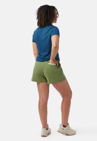 Gröna shorts med sidofickor, blå kortärmad topp och ljusa vandringsskor. Lockigt hår och en avslappnad hållning.