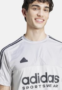 Jersey deportivo gris de Adidas con textura acanalada, franjas de acento negras y un gran logo negro en la parte frontal. Logo bordado en el pecho.