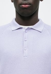 Polo de color lavanda claro con tejido texturizado, que presenta un cuello con solapa y tres botones a juego en la parte delantera.