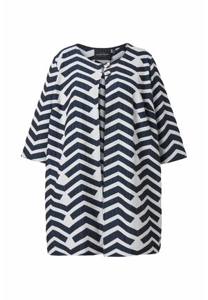 Valkoinen takki, jossa musta siksak-kuvioinen vetoketjukuosi, pyöreä pääntie, puolipitkät hihat ja kaksi nappia yläosan etukappaleessa.