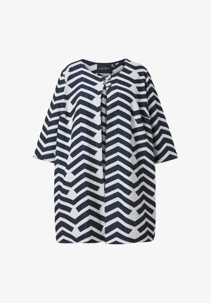 Valkoinen takki, jossa musta siksak-kuvioinen vetoketjukuosi, pyöreä pääntie, puolipitkät hihat ja kaksi nappia yläosan etukappaleessa.