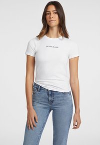 Witte katoenen t-shirt met korte mouwen en ronde hals, met een zwart "GUESS JEANS" logo over de borst, gecombineerd met blauwe jeans.