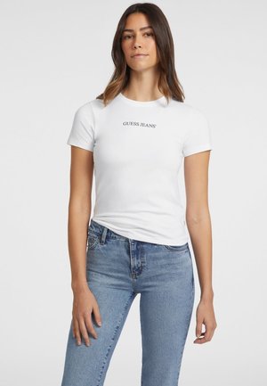 T-shirt en coton blanc à manches courtes et col rond, avec le logo noir "GUESS JEANS" sur la poitrine, associé à un jean bleu.