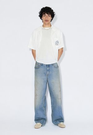 Hvid kortærmet skjorte med knapper, grå t-shirt under, slidte løstsiddende blå jeans og beige fodtøj, med en subtil grafik på skjorten.