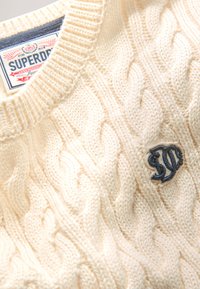 Kabelio rašto kreminis megztinis su apvalia apykakle ir mažym mėlynu siuvinėtu logotipu priekyje. Minkšta tekstūra su storu ribbiniu apykakle.