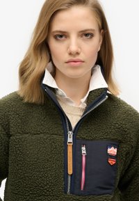 Veste en polaire de couleur vert foncé avec une poche poitrine zippée bleu marine, tirette en cuir et petites nuances. Design texturé, chaud et décontracté.