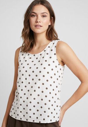 Young woman with shoulder-length brown hair wearing a sleeveless white top with dark polka dots against a plain background.