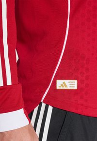 Rød atletisk trøje i mesh stof, med hvide striber langs ærmerne og et guld mærke med logo, sat mod sorte shorts.