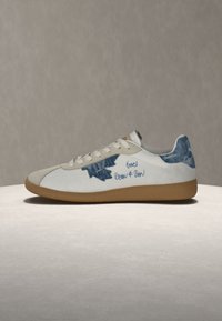 Baskets basses blanches avec bout en daim beige, semelle en gomme, patchs en denim bleu et texte bleu manuscrit sur le côté.