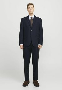 Tumed ülikond ühe nööpauguga bleiseriga, sobivate pükste, valge kleidisärgi ja pruuni lipsuga. Prune suede kingad täiustavad komplekti.