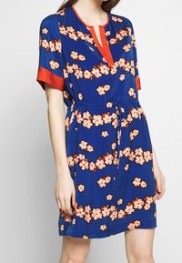 Florale jurk van blauwe stof met roze bloemen, een v-halslijn, korte rode mouwen en een verstelbare tailleband voor een getailleerd silhouet.