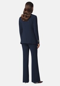 Granatowy, dopasowany garnitur z marynarką o prostym kroju i złotymi guzikami, zestawiony z rozszerzającymi się spodniami. Gładka tekstura tkaniny.