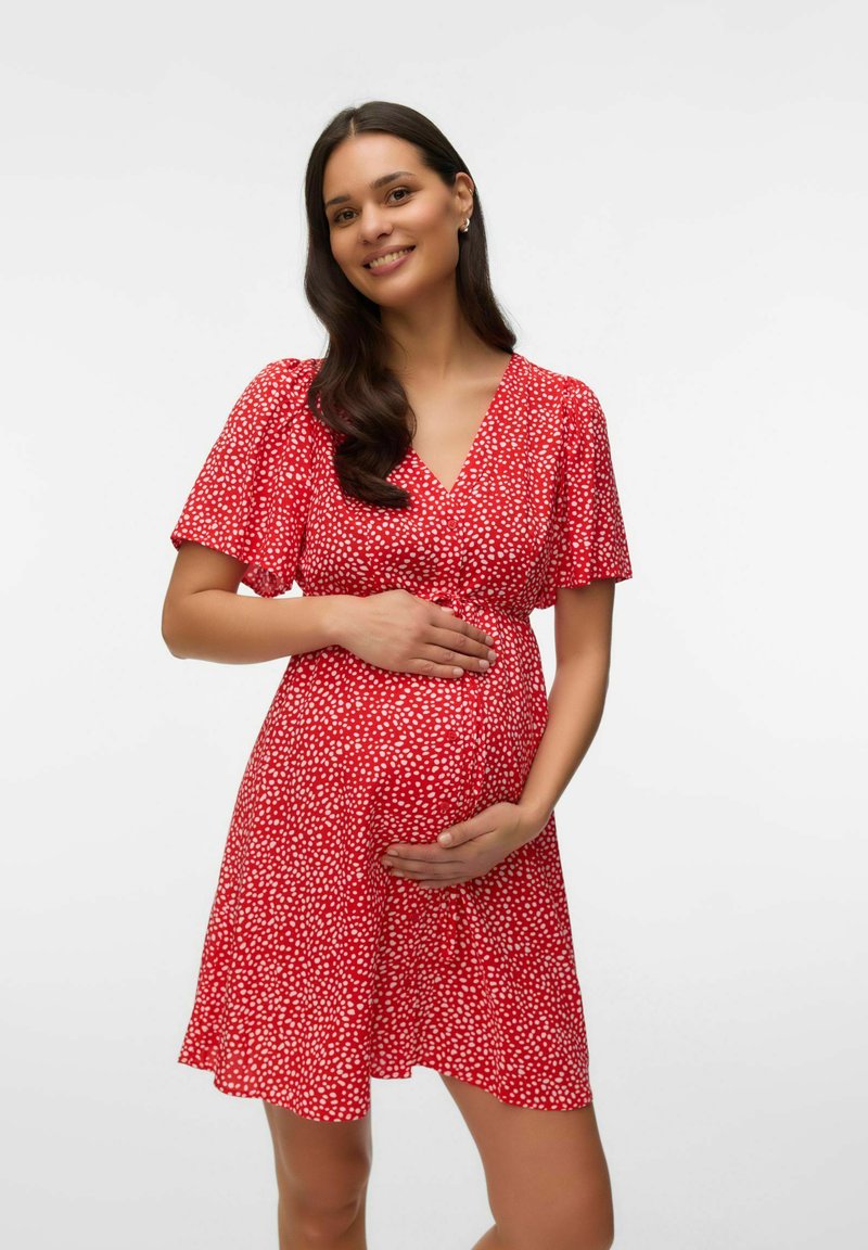 Femme enceinte souriante, portant une robe rouge à pois blancs, les mains caressant doucement son ventre sur un fond uni.