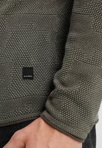 Pull vert tricoté avec un motif texturé, comportant une étiquette noire GABBA sur l'ourlet et une coupe ajustée au niveau des poignets.