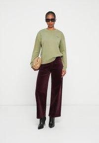 Sudadera verde oliva de manga larga y cuello redondo, combinada con pantalones de pana burdeos de pierna ancha y botas negras de tacón. Sosteniendo un bolso beige.