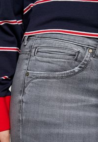 Szare dżinsy o gładkiej fakturze, z przednią kieszenią i widocznym szwem. W zestawie z ciemną koszulą z długim rękawem w czerwono-białe paski.