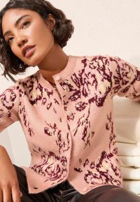 Rozā kardigans ar ziedu rakstu bordo un dzeltenā krāsā, ar apaļu kakla izgriezumu, pogu aizdari un mīkstu, teksturētu adījuma audumu.